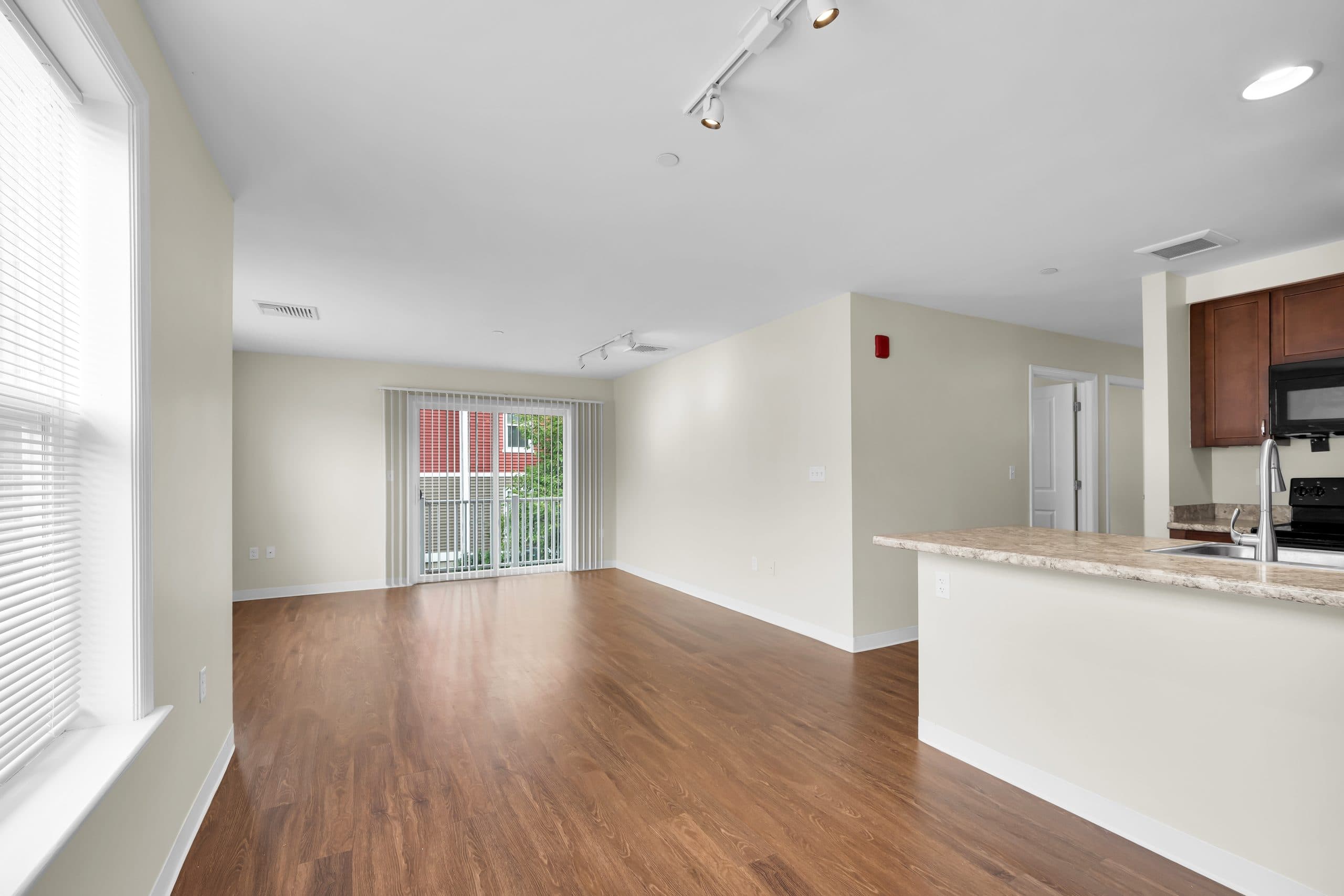An open living room with plank flooring, a breakfast bar into the kitchen, and a sliding door to a balcony